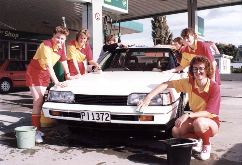 Five young women in bright yellow and red football uniforms are grouped around a white car. They are washing the car, probably at a petrol station. 