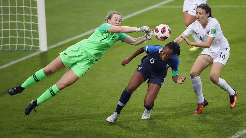 A young woman in green New Zealand goalkeeper uniform leaps into the air while reaching to push a ball away from a goal during a football game. A woman in a blue football uniform is partly crouched in front of her to avoid the punch, while other players in white New Zealand uniforms run towards her.  