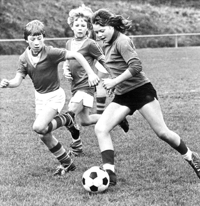 A teenage girl in football uniform with a black and white football at her feet, being chased by two teenage boys in a different football uniform during a football game. 