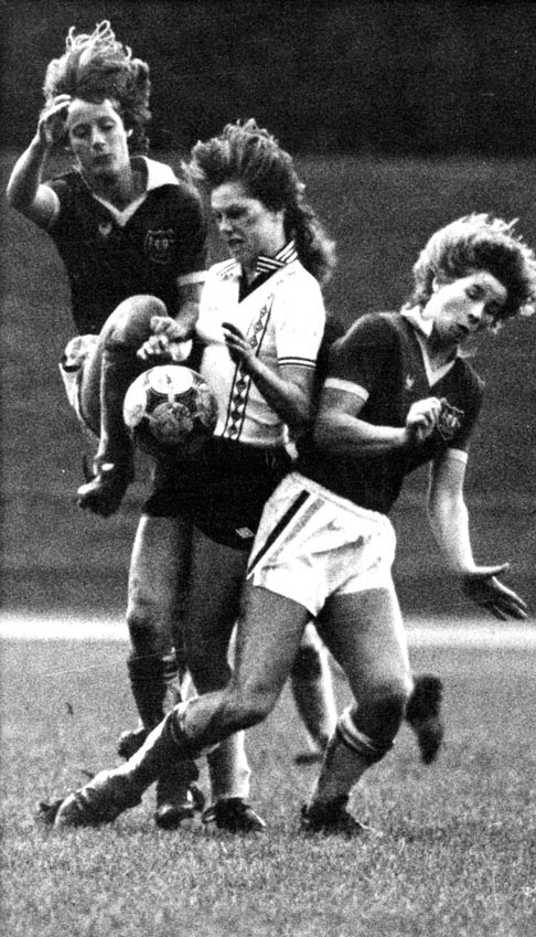 A woman in a New Zealand football uniform, with a football at hip level, sandwiched between two players in Australian football uniforms, one of whom has jumped up in the air. 