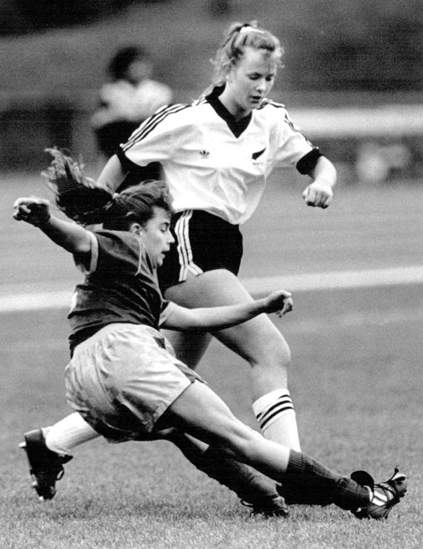 A young adult girl in a white New Zealand football uniform with a black fern symbol, during a football game, with a young adult girl in Australian uniform appears falling to the ground.