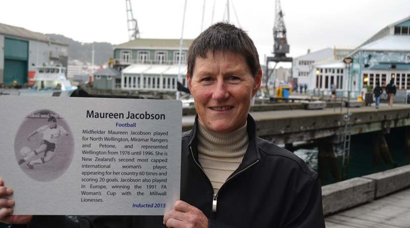 A woman in her 50s stands on the Wellington waterfront holding a white plaque which contains a photo of her playing football as a younger woman, and text about her career.