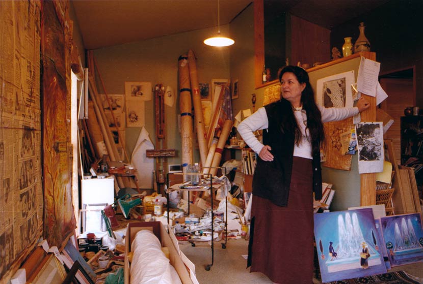 Pauline Thompson, in middle age, stands in a room cluttered with artworks, cardboard storage rolls, paints and other artists’ equipment. 