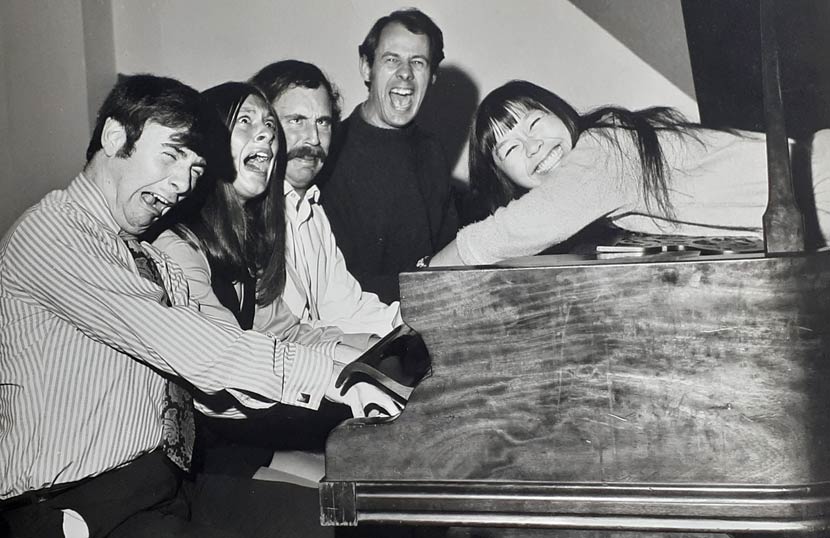 Four people seated at a piano and one lying on top, all pulling faces. 