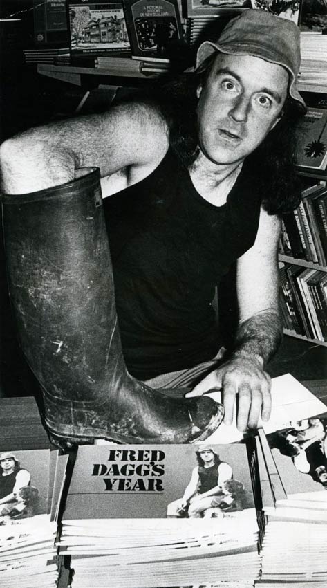 John Clarke, dressed as Fred Dagg in a black singlet and floppy hat, sits at a table with a pile of copies of the book Fred Dagg’s year in front of him. His arm is inside a gumboot.