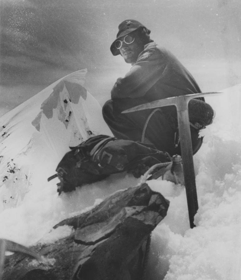 A man crouches on a snowy mountain peak with a bag and a pickaxe.