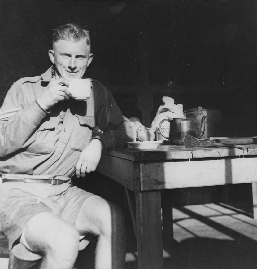 A man in military uniform sits at a wooden table and drinks a cup of tea.