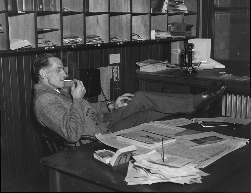 A seated man lights a cigarette, his feet on his cluttered desk, in front of shelves filled with papers. 