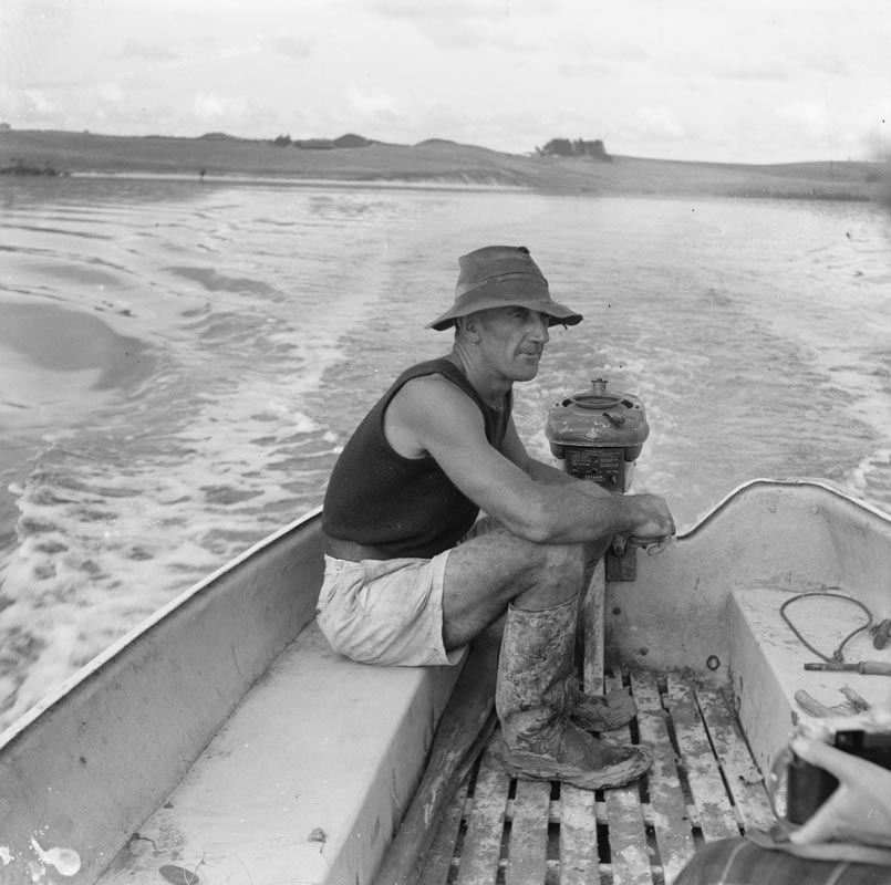 A man in a black singlet and floppy hat seated in a motorboat in a large body of water, the wake rippling out behind the boat.