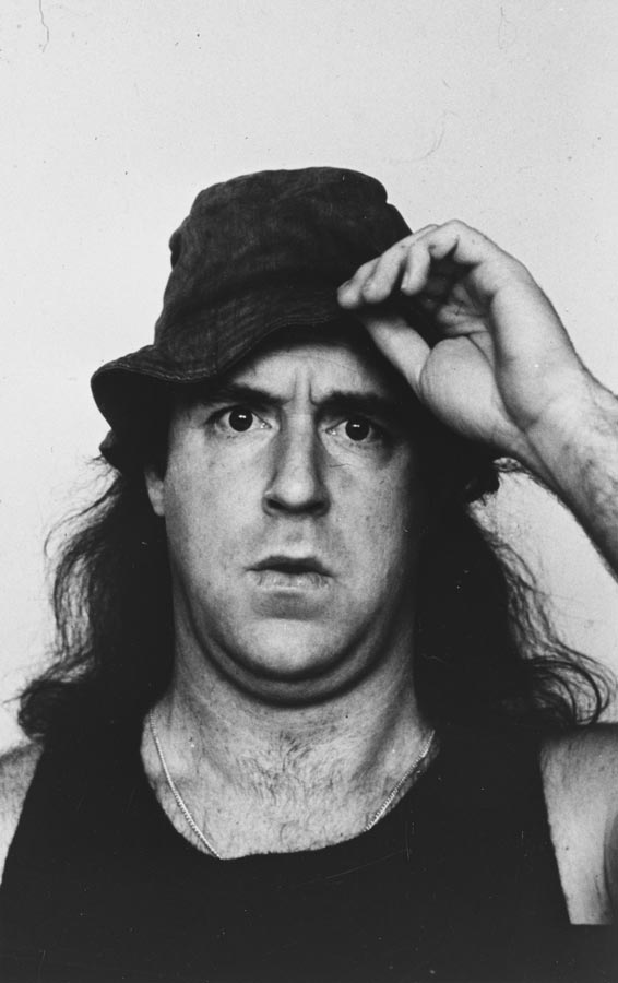 A man with long hair, a black singlet, and a hat, looking bewildered.