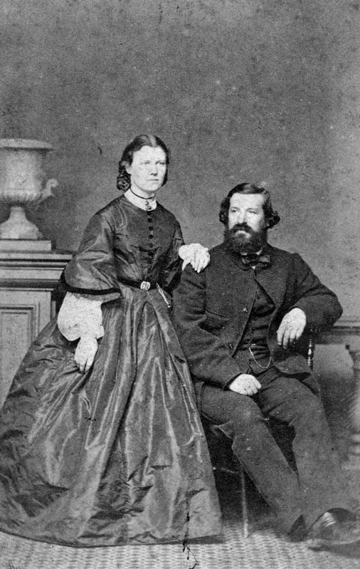 A studio portrait in which a standing woman, in a full-length Victorian dress, leans against a seated, bearded man in a suit.