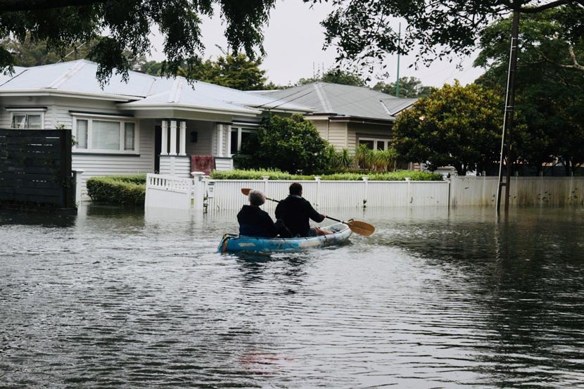 Two people travel down a flooded suburban street in a kayak.