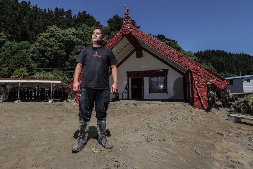 A man stands on a large pile of silt in front of a wharenui. 