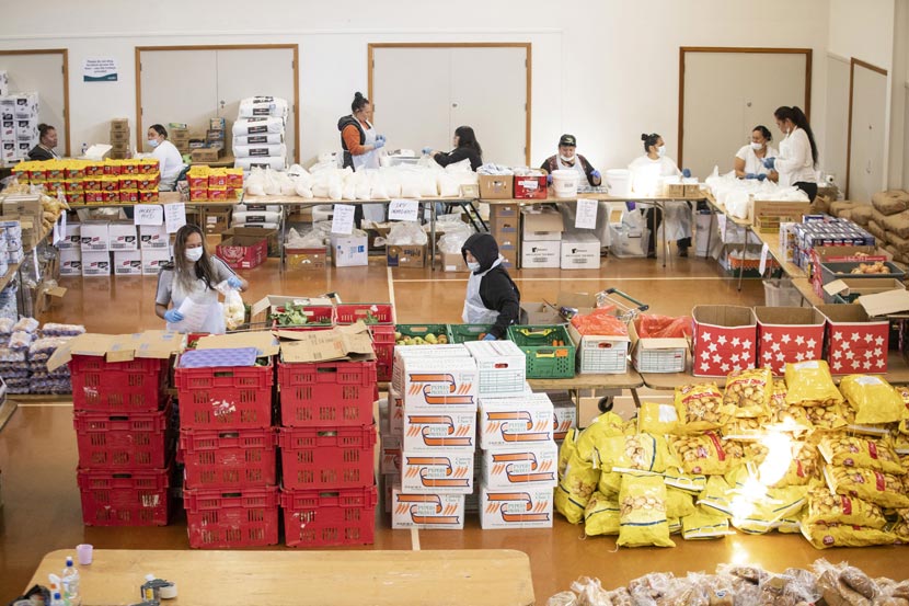 A group of people wearing masks, in a large hall, surrounded by many boxes and bags of food.