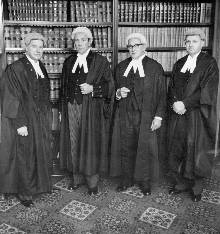 Four judges, in judicial wigs and gowns, standing in front of shelves of law books.