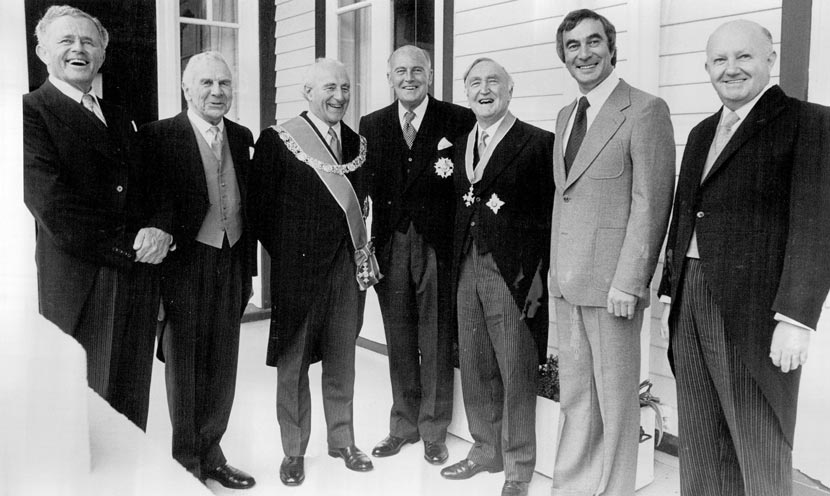 Seven men in suits, some with honours pinned to their jackets.