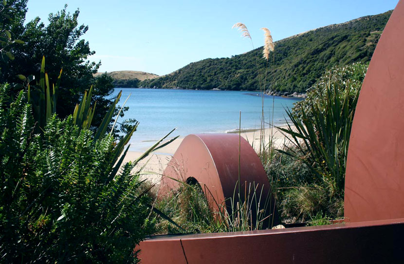 Red steel sculpture set amongst trees and bush with golden sand beach and sea in the background