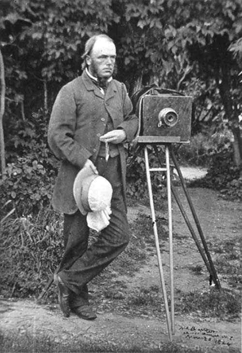 An outdoors photograph of Alfred Barker standing beside a camera on a tripod 