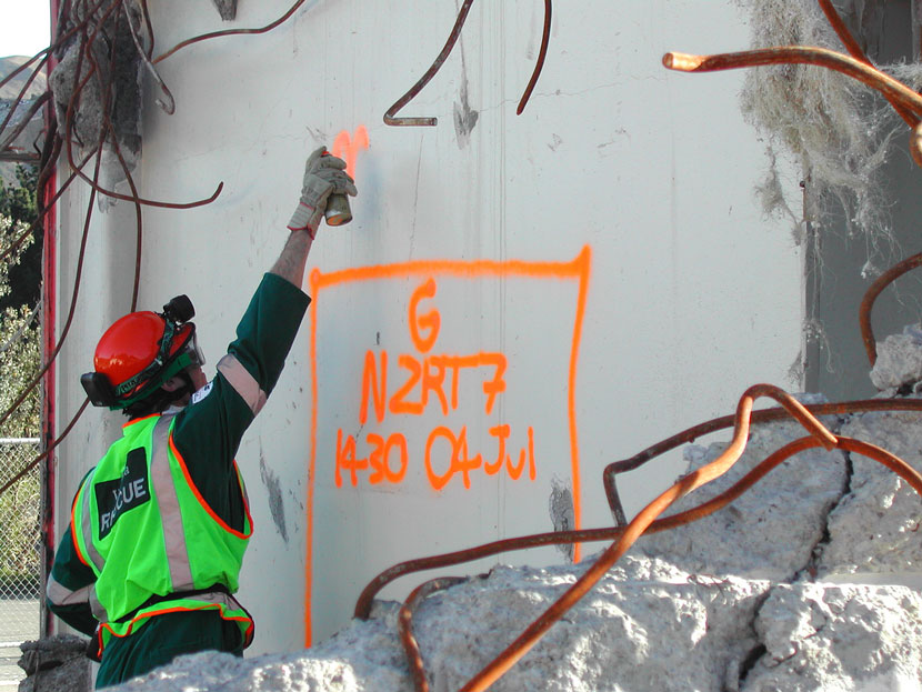 A person in safety gear spraypainting words and numbers on a wall surrounded by building wreckage. 