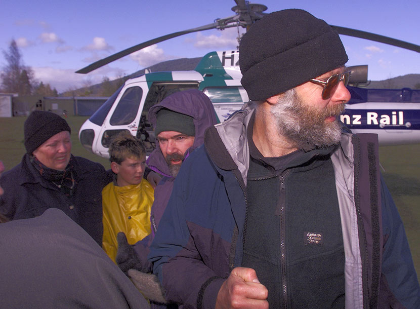 A man in a woolly hat stands on front of a couple and their child, and a helicopter. 