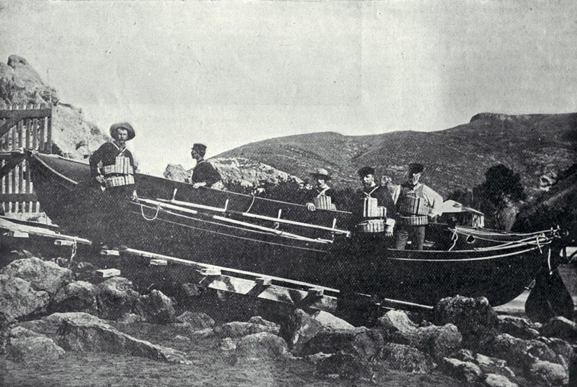 Long rowing boat pulled up on rocks surrounded by five men wearing life jackets. 