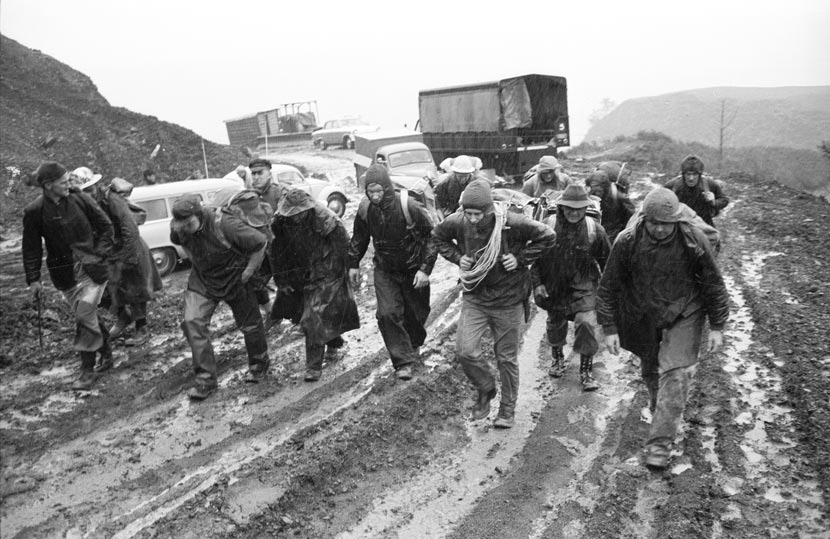 A large group of people in wet weather clothing set off up a muddy road with a truck behind them. 
