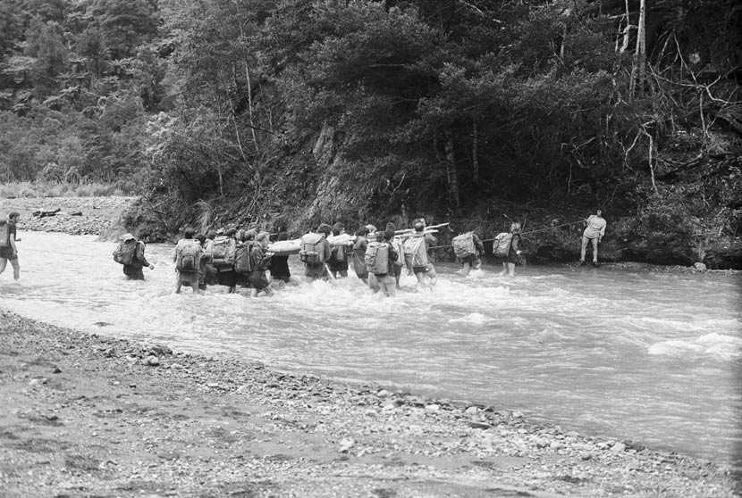 A large group of people with tramping packs walk in a river, some carrying a stretcher 