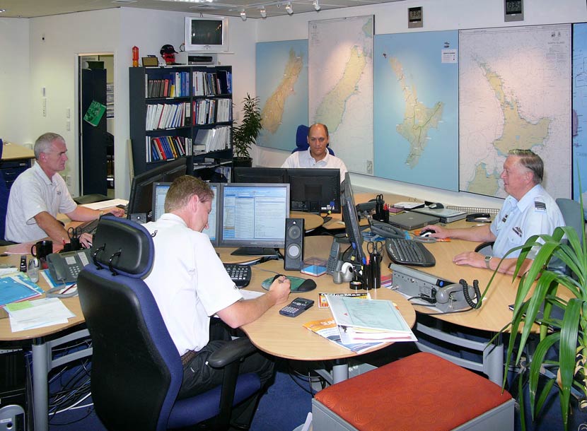 Four men sit in front of computers with large maps displayed behind them 