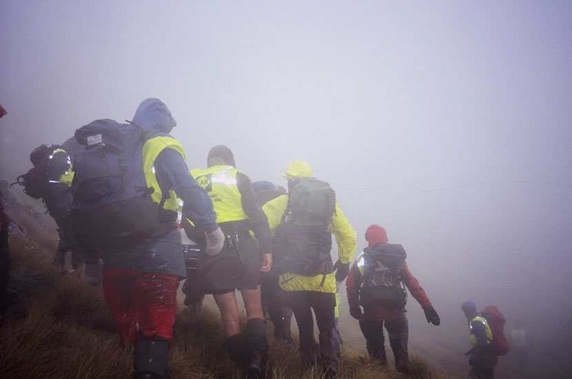A large group of people in tramping clothing walk on a steep hill in fog. 