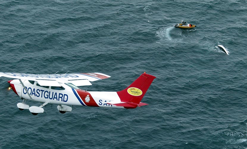 A small plane labelled ‘Coastguard’ flies above a rescue boat and an overturned boat in the sea 