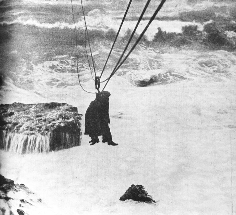 Two people suspended on wires high above a churning sea and rocks