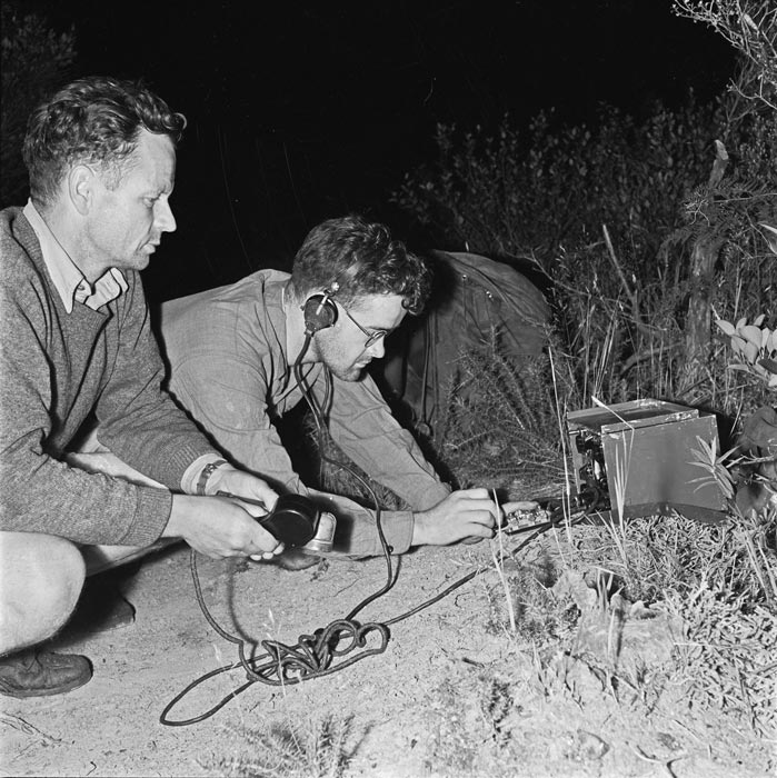 Two men seated in the bush in front of a radio and wires.