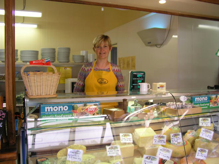 Woman in yellow apron standing behind cabinet filled with different cheeses.