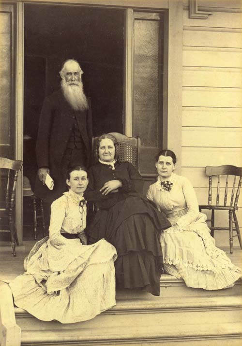 Leonard Williams in later life, with a long white beard, standing on a verandah beside Sarah Williams and their two adult daughters. 