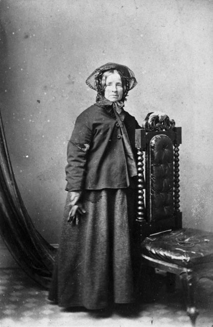 Eliza Wohlers in a hat, scarf, coat and gloves, standing beside a chair in a photographer's studio.. 