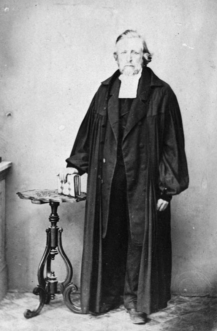 Johann Wohlers in clerical garb standing beside a table and holding a book.