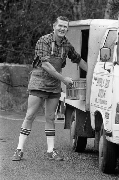 A milkman in shorts and socks, taking milk from his truck.