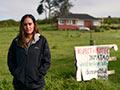 Pania Newton standing in green field with protest sign and house in the background.
