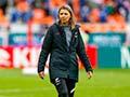 A woman in black coat with a white fern symbol walks on a football field, with a player out of focus in the foreground.