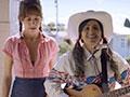 Two women, on holding a guitar and harmonica, standing together dressed in country western clothes.