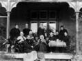 A group of well-dressed middle aged and elderly Pākehā standing and sitting on the verandah of a house beside a table with a teapot and cups.