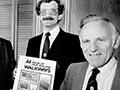 Three men in suits stand side by side, smiling at the camera, and holding a copy of the AA book of New Zealand walkways.