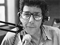 A photograph of Paul Holmes as a young man, wearing headphones and seated in front of a microphone.