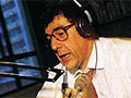 Paul Holmes, wearing headphones and speaking into a microphone, seated at a studio console.