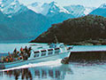 A colour image showing a body of water surrounded by forested and snow-capped mountains, with a motor vessel in the centre, heavily laden with passengers. Two people picnic in the right foreground. 