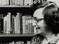 The photograph shows Ruth Dallas, in middle age, standing in profile beside a shelf of books, and holding a book open in front of her. 