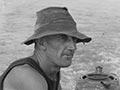 A man in a black singlet and floppy hat seated in a motorboat in a large body of water, the wake rippling out behind the boat.