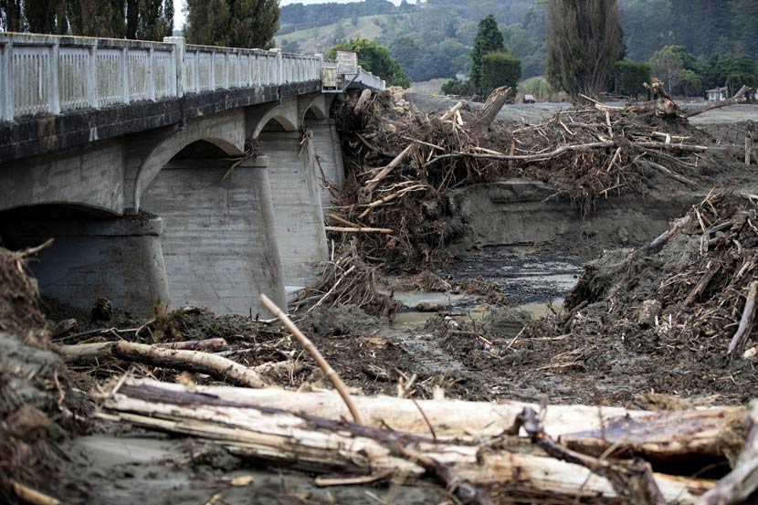 Large trees and other debris lying up against a low concrete road bridge across a river.