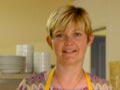 Woman in yellow apron standing behind cabinet filled with different cheeses.