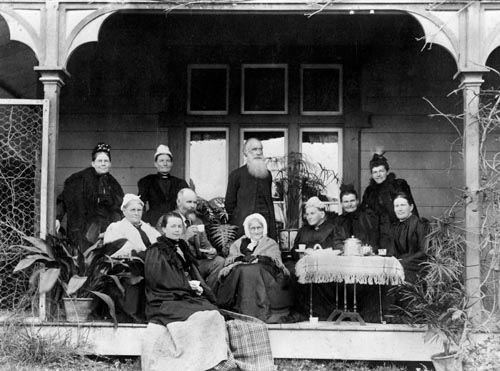 A group of well-dressed middle aged and elderly Pākehā standing and sitting on the verandah of a house beside a table with a teapot and cups.